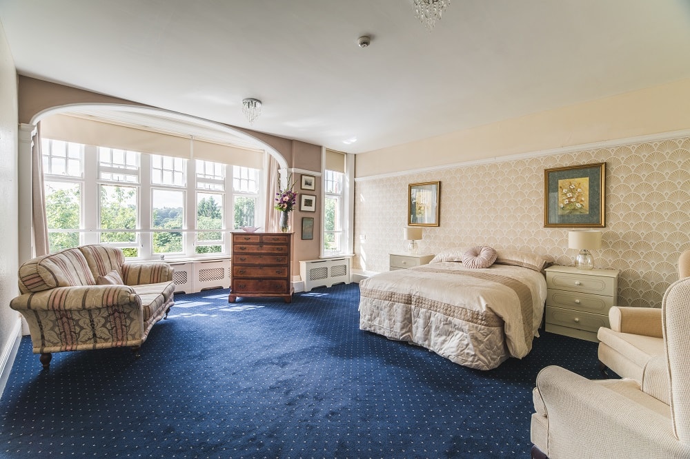 Bedroom interior with a comfortable double bed, blue carpet, fan wallpaper, soft lighting, and large windows overlooking a green landscape.