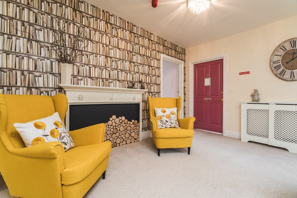 Living room featuring two yellow armchairs with decorative pillows, a fireplace with log storage, book-themed wallpaper, red door, and a large clock on the wall.