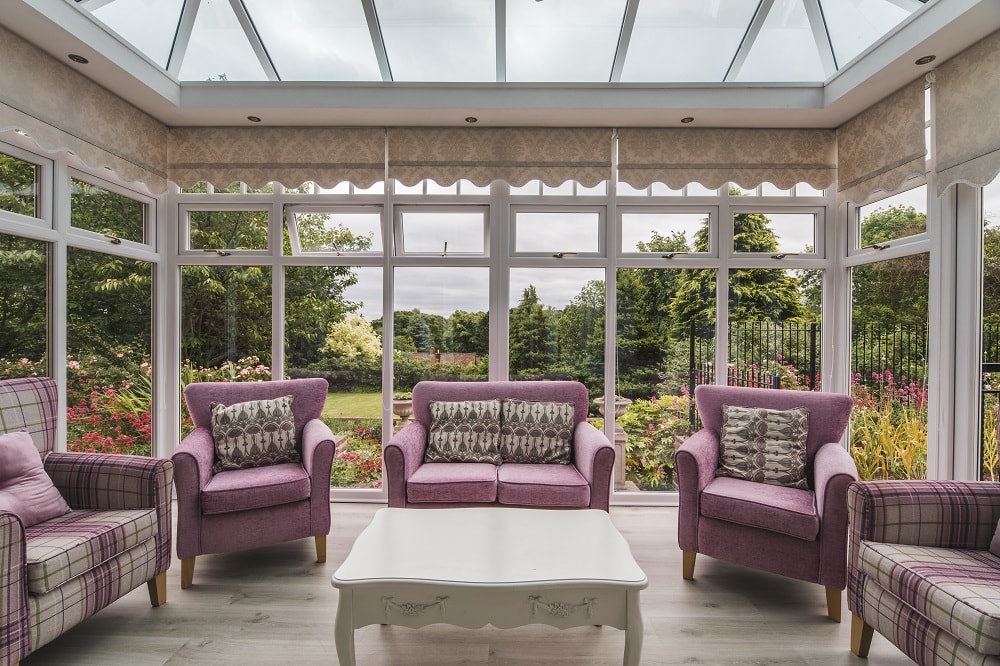 Bright sunroom with large windows and glass ceiling featuring pink sofas and armchairs around a white coffee table with a scenic view of greenery outside.
