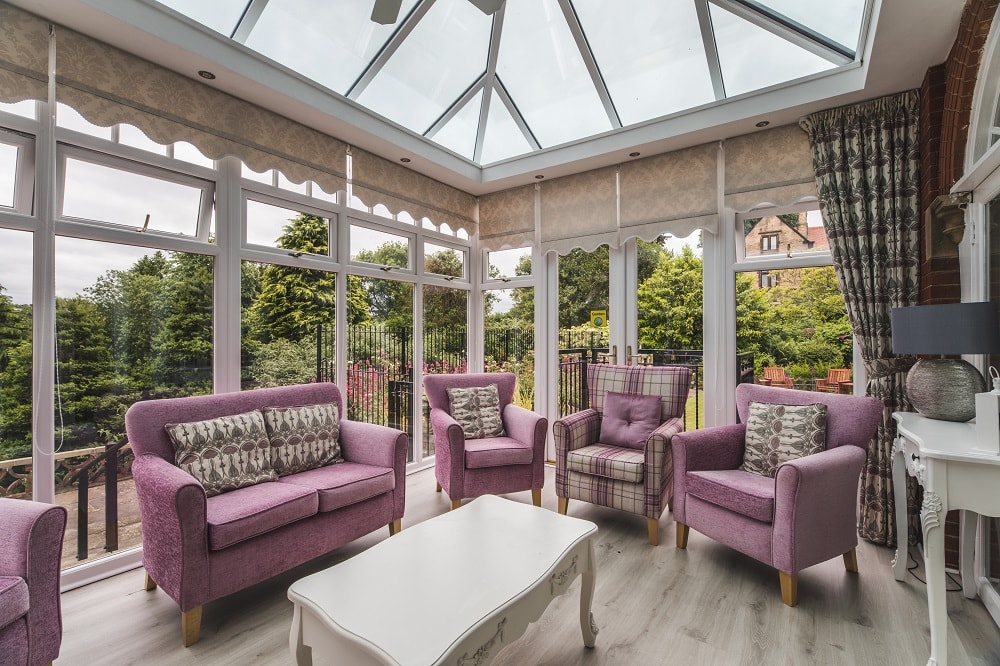 Bright sunroom with large windows and glass ceiling featuring pink sofas and armchairs around a white coffee table with a scenic view of greenery outside.