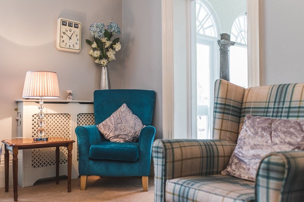 Living room with a teal armchair and plaid wingback chair. A table lamp, decorative vase, and wall clock are decorating the room.
