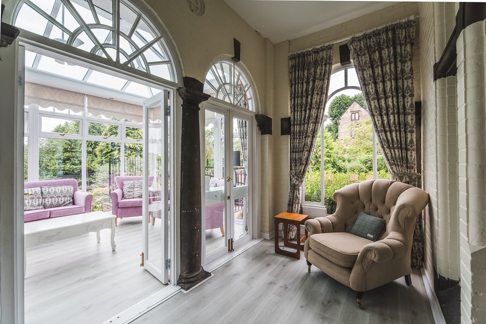 Sunlit room with elegant arched windows overlooking a lush garden with beige armchair and purple sofas