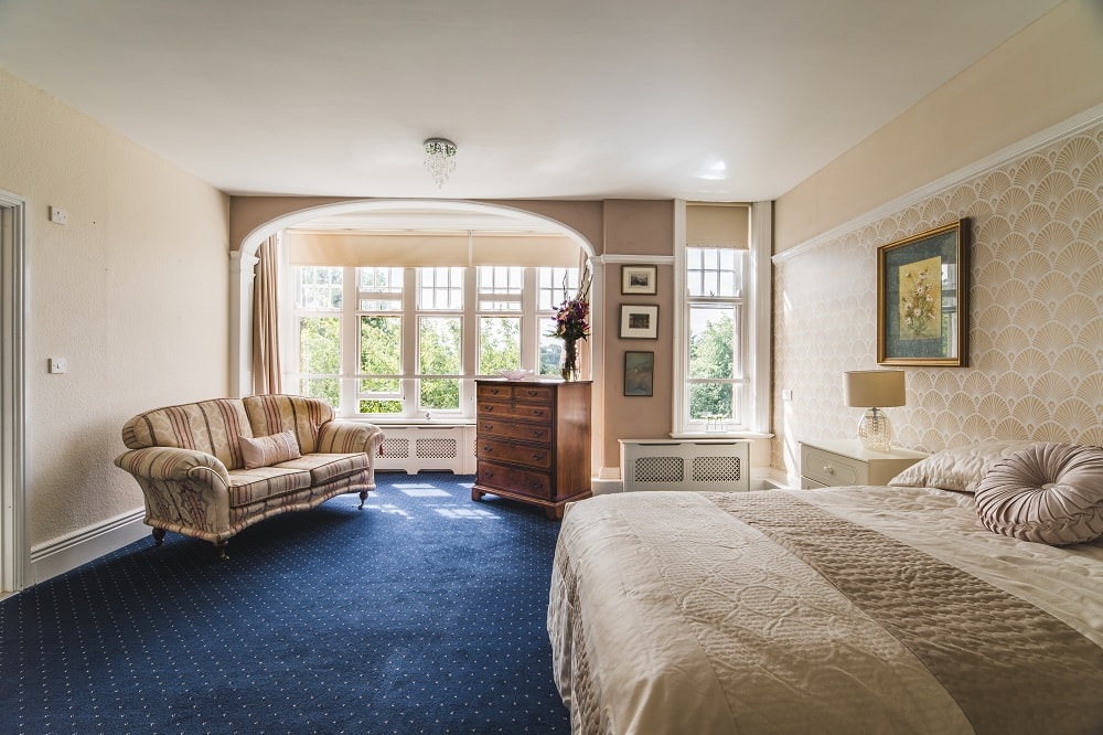 Bedroom interior with a comfortable double bed, blue carpet, fan wallpaper, soft lighting, and large windows overlooking a green landscape.