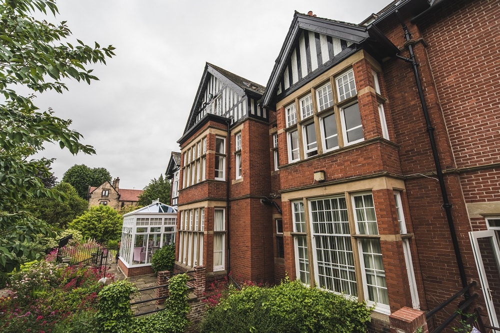 Red brick Tudor-style building with black and white gables surrounded by lush greenery on a cloudy day