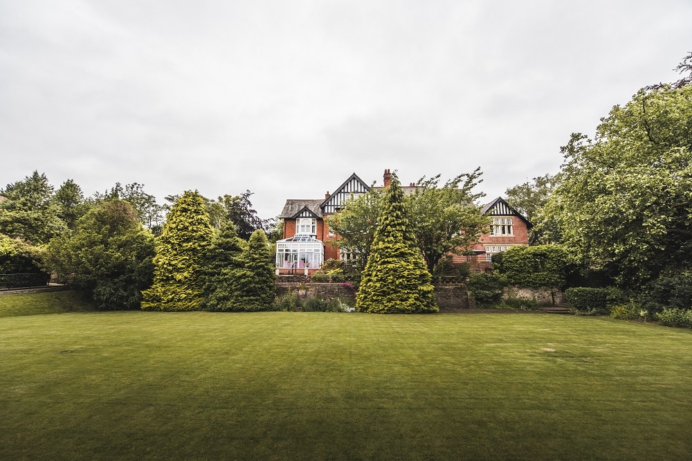 Foreground view of rich green grass framed with lush green shrubbery with a red brick building in the background