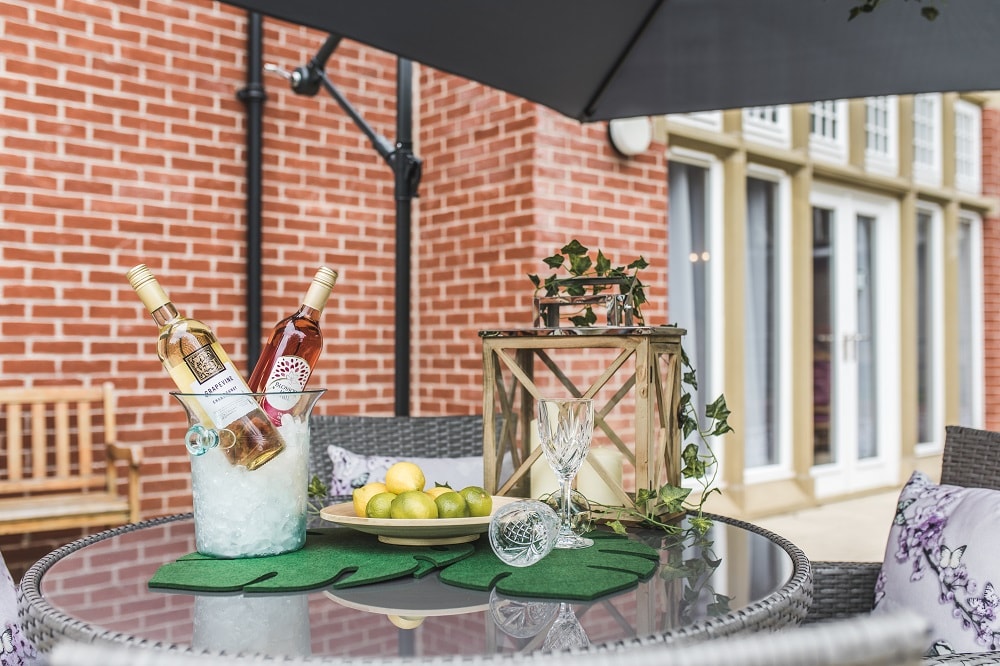 Glass table with bottles of wine in a wine cooler with fruit in a platter and glasses, and a brick building in the background.
