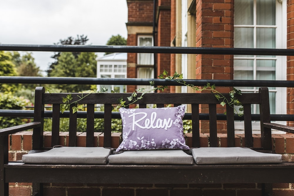 Wooden outdoor bench with a cushion featuring the word "Relax" placed on it surrounded by greenery and a brick building in the background.