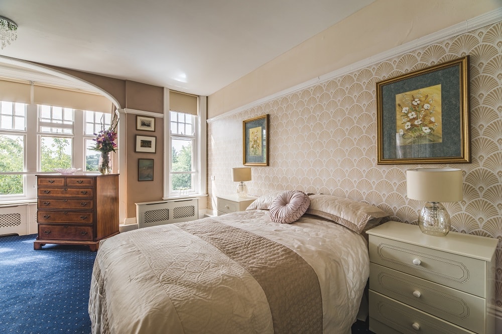 Bedroom interior with a comfortable double bed, fan wallpaper, soft lighting, and large windows overlooking a green landscape.