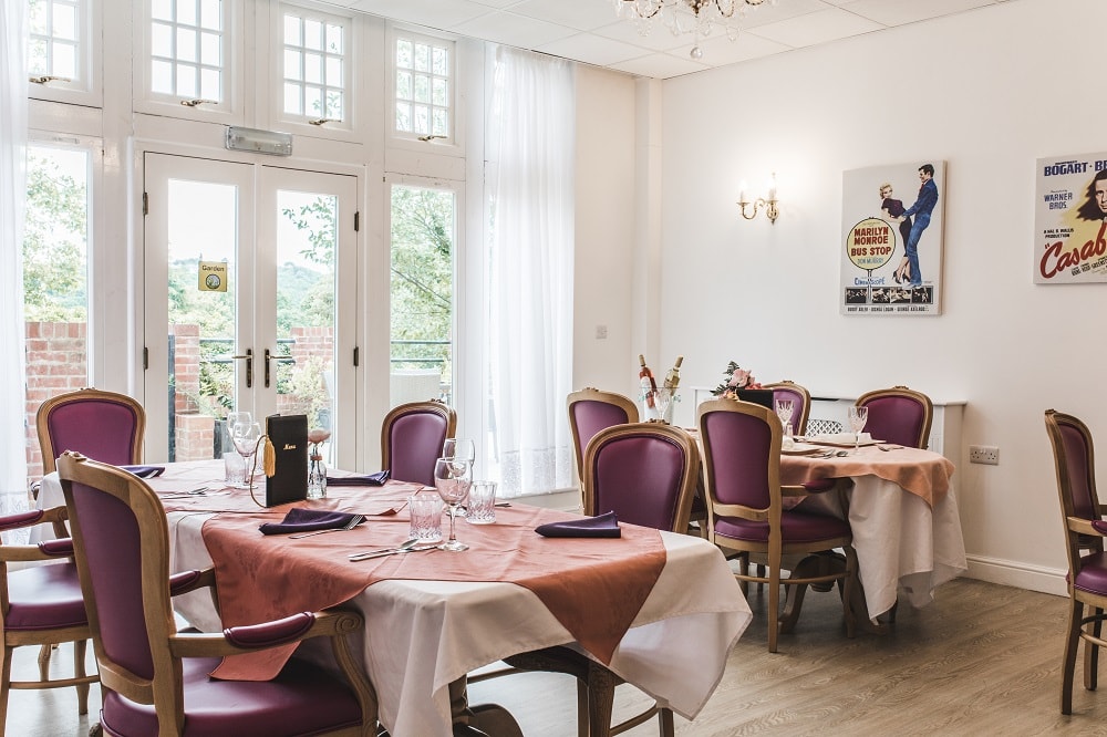 Dining room with tables covered in pink and white tablecloths set for guests Purple chairs surround tables near large windows
