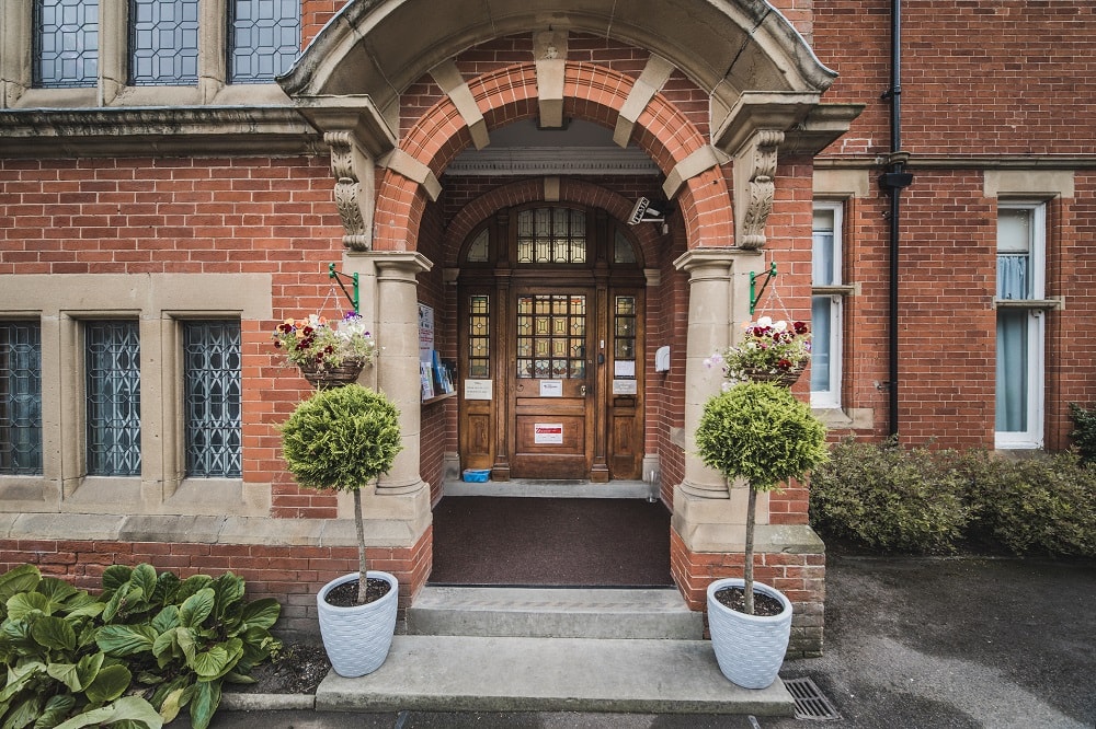 Brick building entrance with arched doorway framed by two potted topiary plants decorative windows on the side and a paved pathway leading to the door.