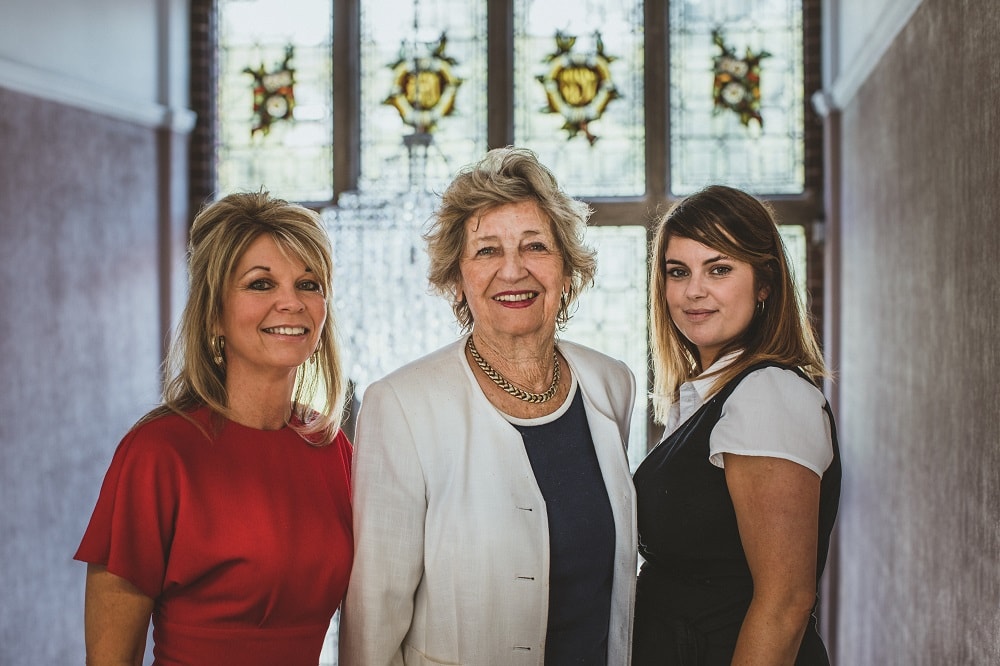 Three women standing together indoors in front of a decorative window smiling and dressed in professional attire