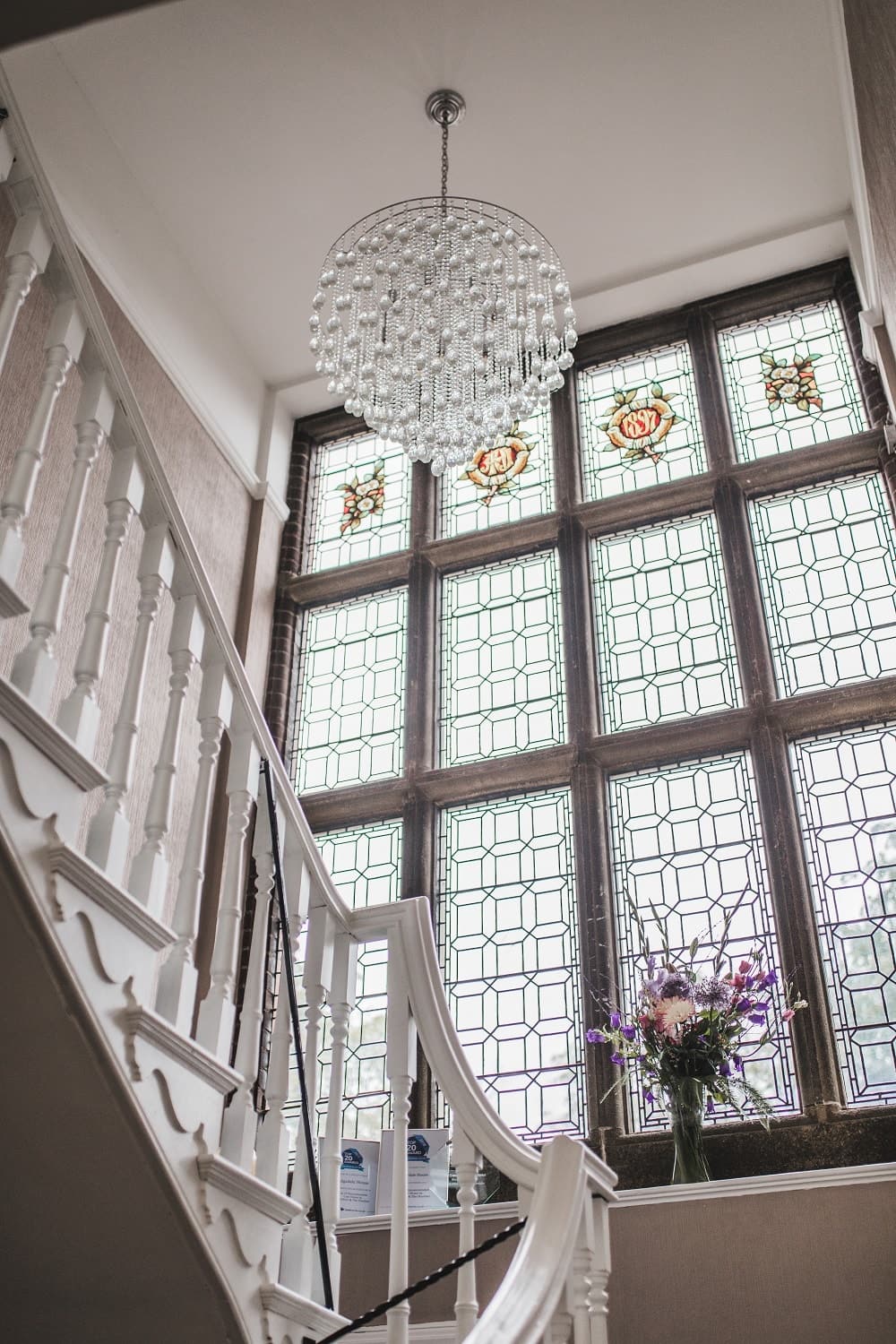 Elegant staircase with white banister and decorative stained glass window backdrop illuminated by a crystal chandelier. A vase of purple flowers adds a charming touch.