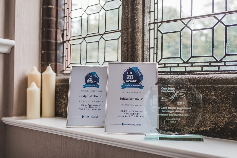 Award certificates and a glass plaque displayed on a windowsill next to candles in a well-lit room