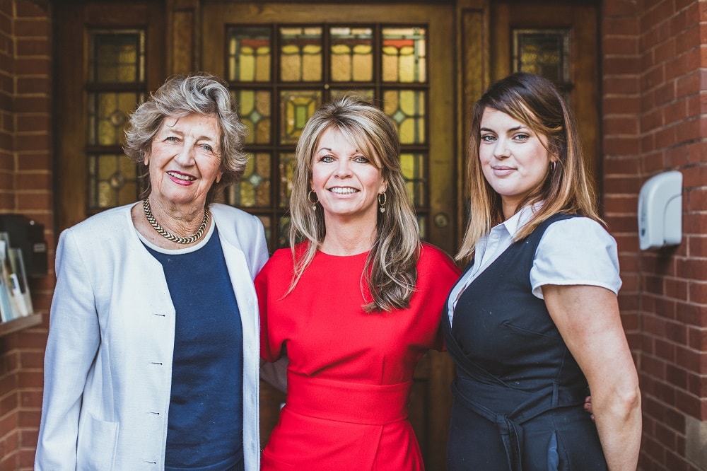 Three women smiling while standing in front of a wooden door with stained glass detail